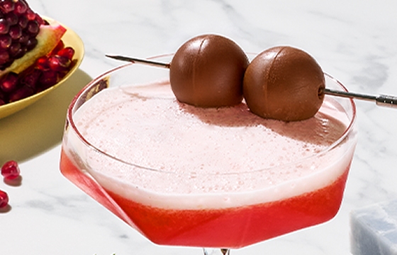 Two bah ham bug sours in geometrical glasses, one cocktail is garnished with 2 Lindt chocolate balls, with a plate of pomegranate seeds to the side